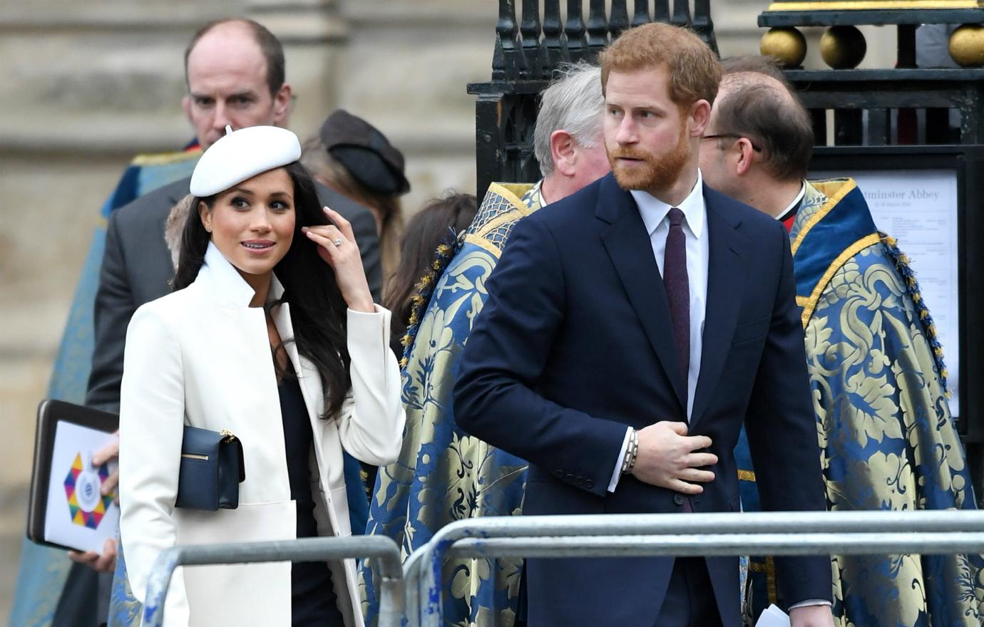 Meghan Markle, in a white beret, white coat and black dress walks next to Prince Harry in a blue suit.