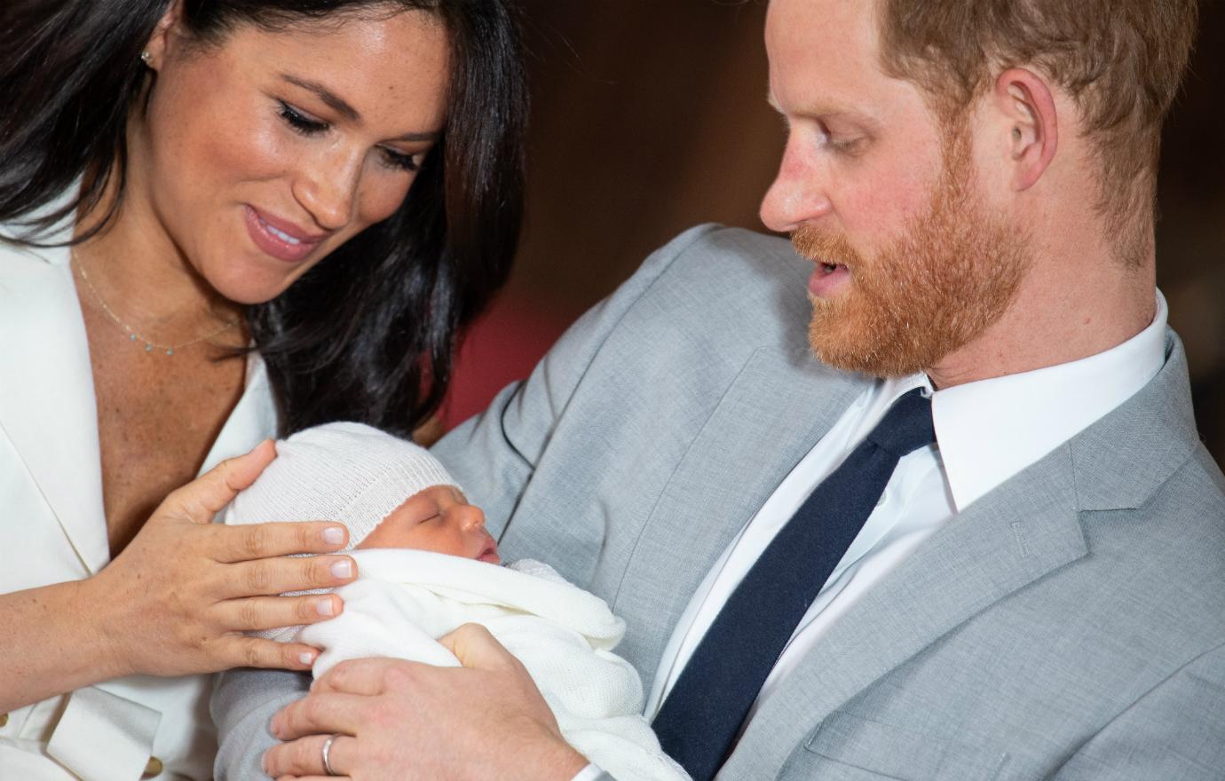 Meghan Markle, in a white dress, cups the head of baby Archie who is swaddled in white clothes and held in the loving arms of Prince Harry who wears a light grey suit and white shirt.