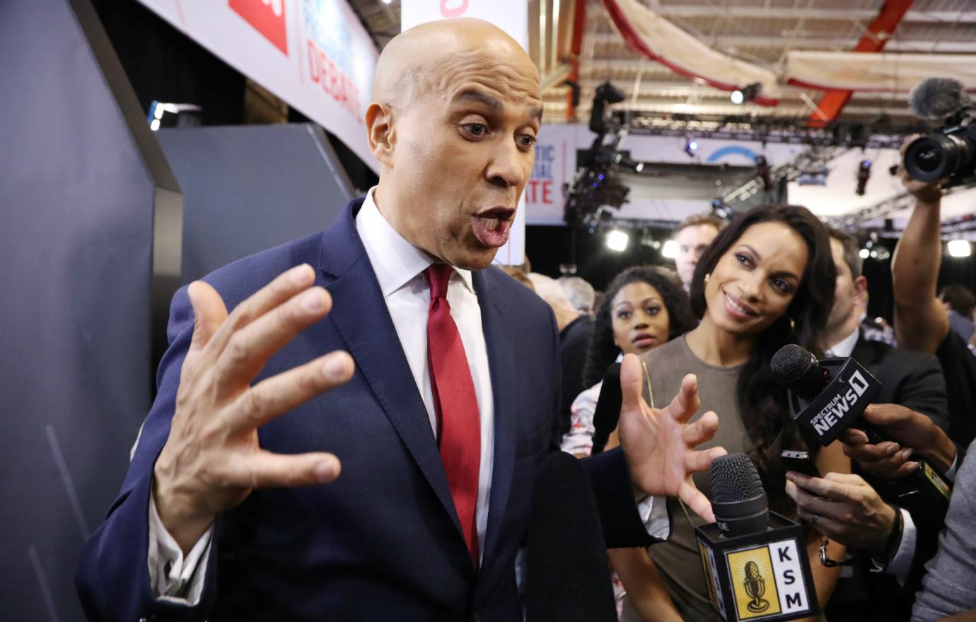 Cory Booker, in suit and red tie, gives an interview while a beaming Rosario Dawson, in a lime green dress, looks on.