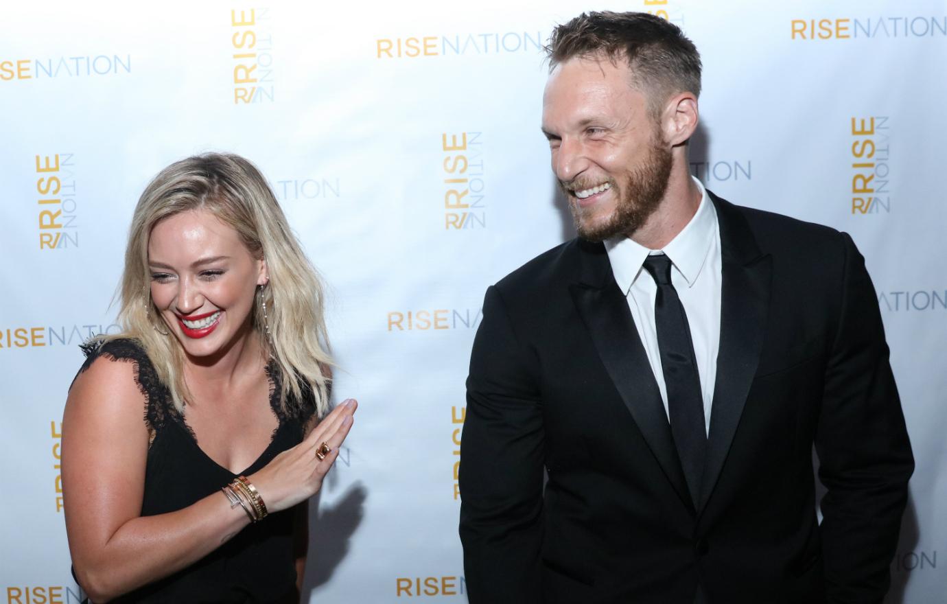 Hilary Duff, in a black dress, stands next to Jason Walsh who wears a black suit and white tie on a red carpet.