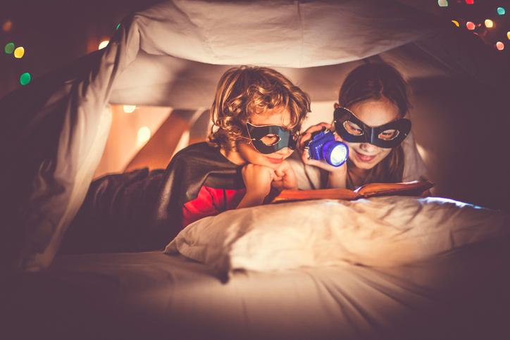 Two children are reading a book in bed under a sheet. Little boy is wearing a superhero costume. They are hiding under a homemade playing tent, made of bedding. Next to him, the little girl is reading a story book with a flashlight in her hand