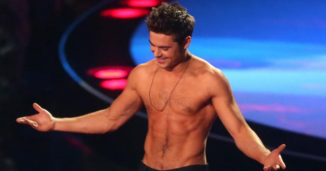 A shirtless Zac Efron stands on a stage with his arms widespread.