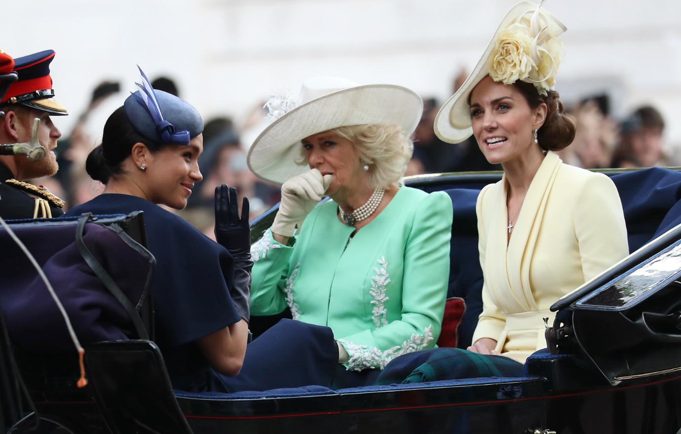 Meghan sits in a royal carriage. She wears a dark blue dress. Sitting opposite is Kate Middleton in a yellow dress and hat and Camilla who wears a mint green dress and a white hat.