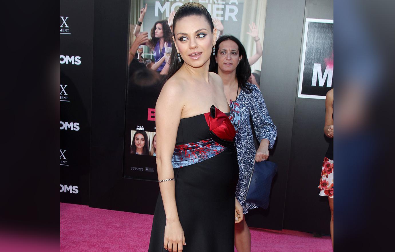 A pregnant Mila Kunis stands on the red carpet in a black dress with a red sash belt.