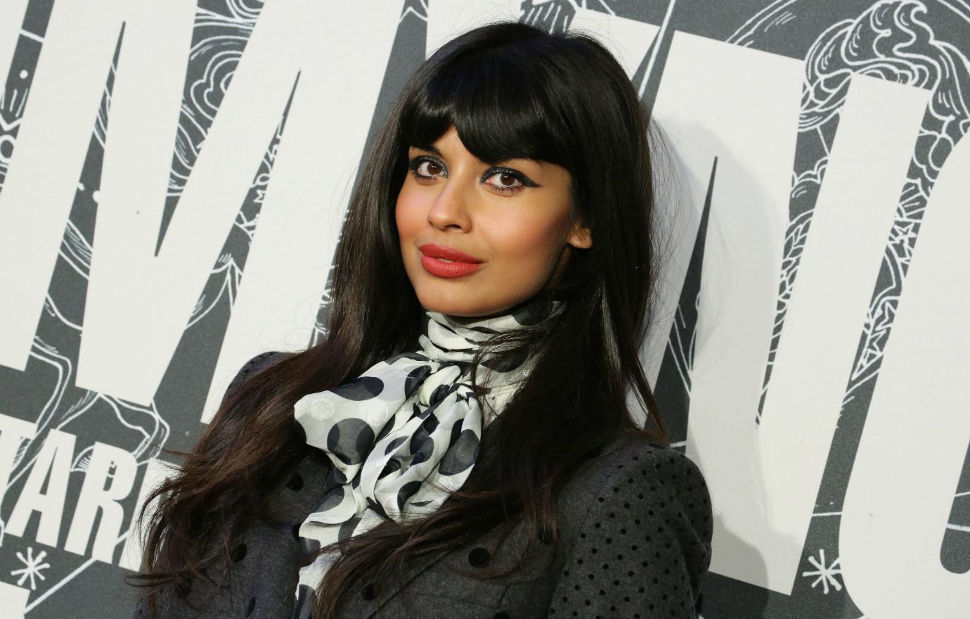 Jameela Jamil in a black and white scarf and black jacket stands on the red carpet.