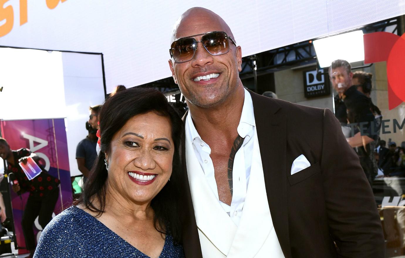 The Rock, in a white shirt and jacket, smiles alongside his mom on the red carpet.