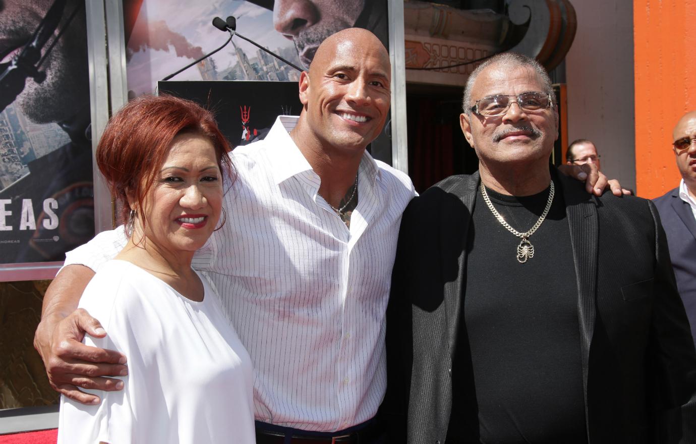 The Rock, in a white shirt, stands on the red carpet flanked by his parents.