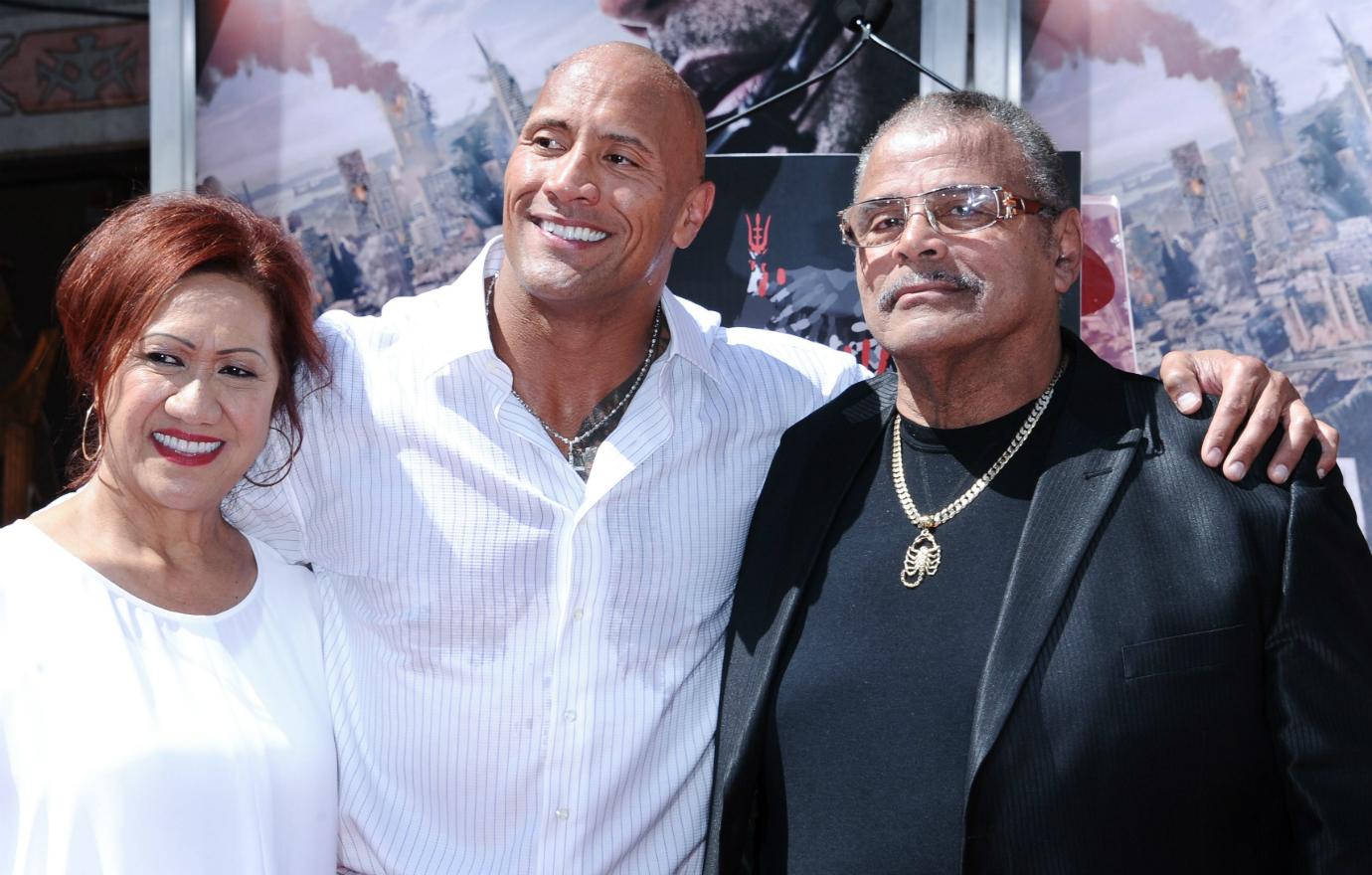 The Rock, in a white shirt, stands on the red carpet flanked by his parents.