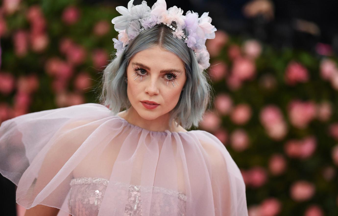 Lucy Boynton in a grey pink dress with a tiara of flowers on her head.