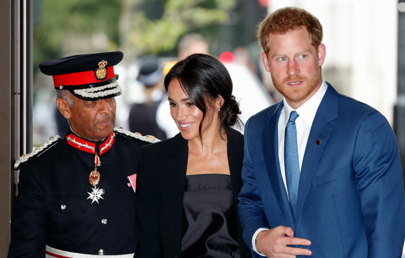 Meghan Markle, in a black outfit and clutch, walks with Prince Harry in a blue suit and white shirt past a man decked out in a royal military costume.