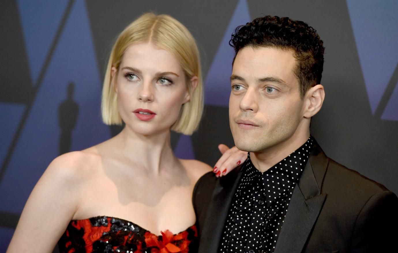 Lucy Boynton, in a red and black dress, poses with Rami Malek on the red carpet. He wears all black.