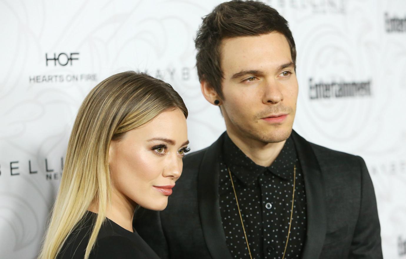 Matt Koma in a black jacket and black shirt with white spots poses on the red carpet next to Hilary Duff who wears a black dress.