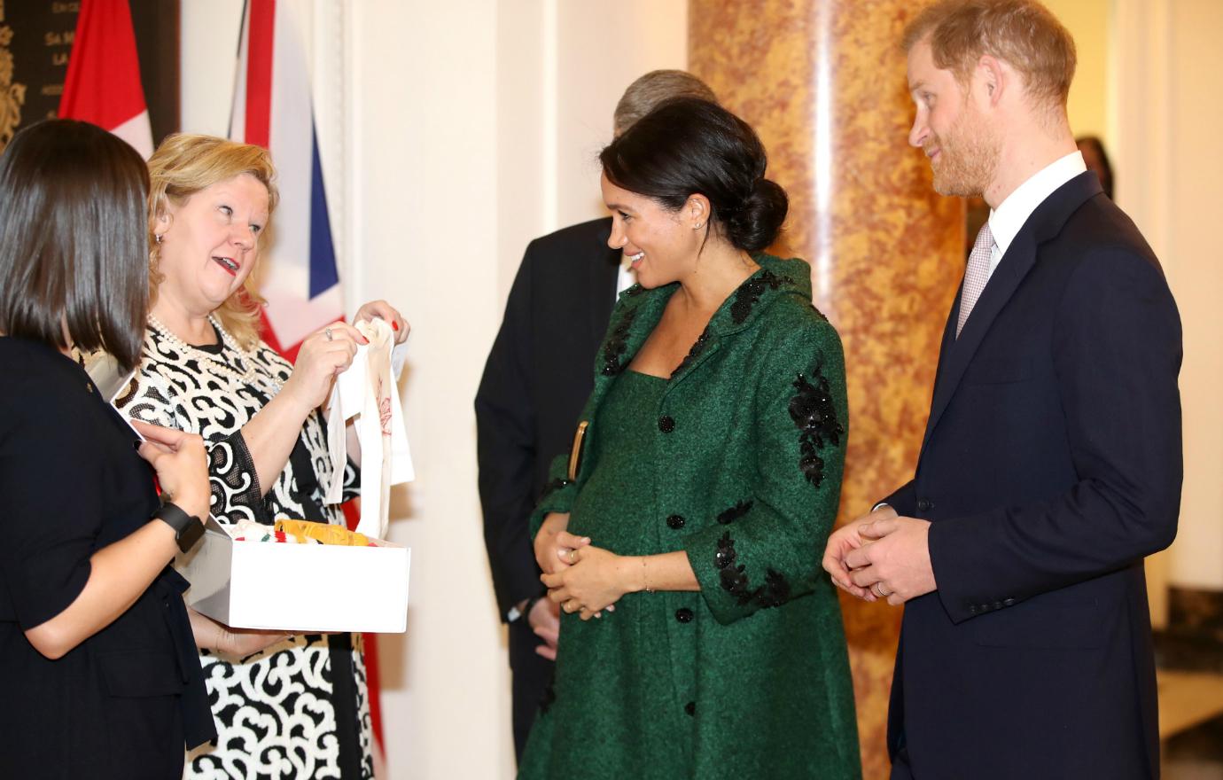 Meghan, wearing a green dress and coat, and Prince Harry, in a dark suit and white shirt, admire a baby onesie that a woman is holding up for them to look at.