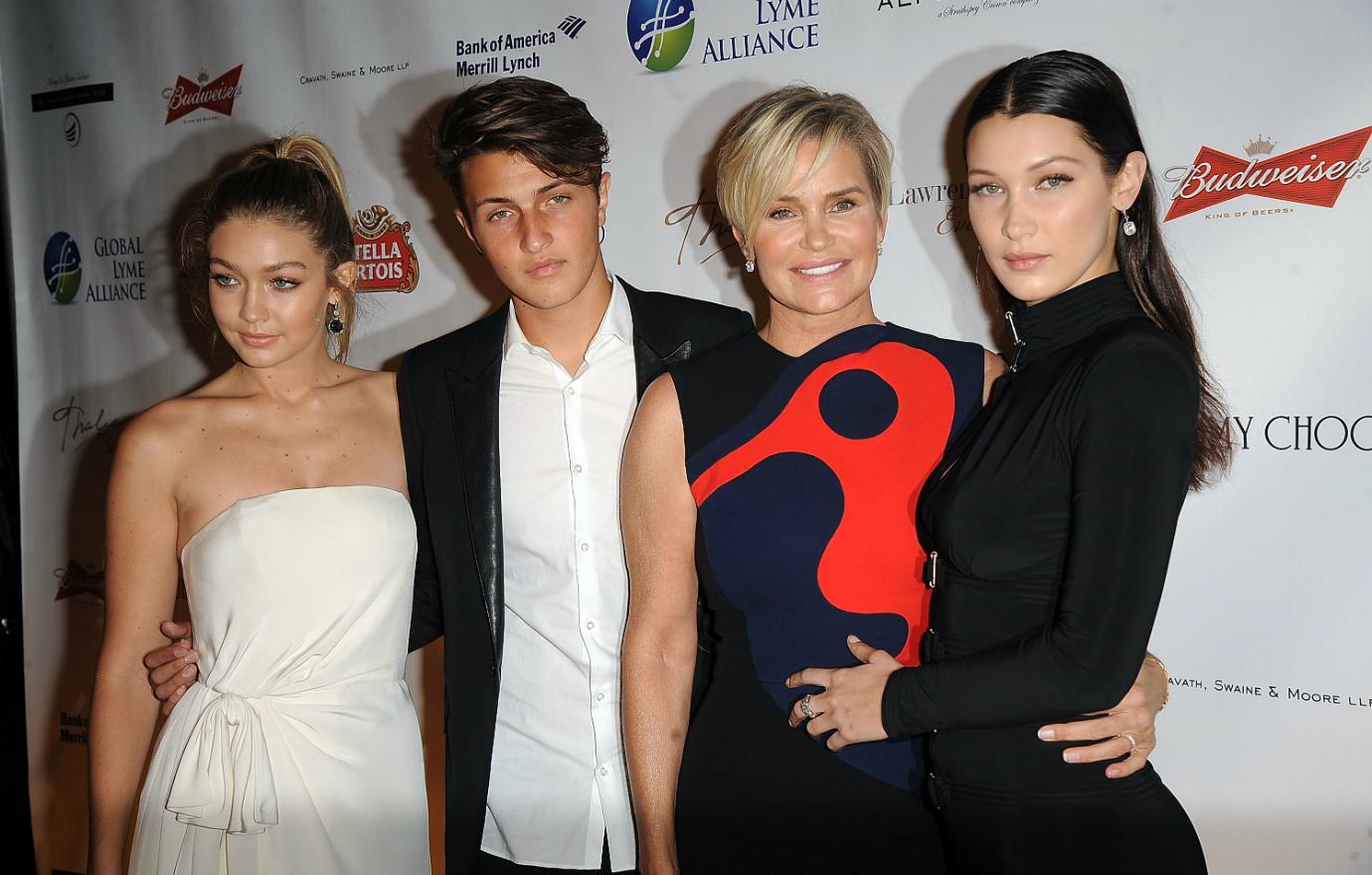 Yolanda Hadid wears a blue and black sleeveless dress with a large red design, posing with her kids, Gigi, Anwar, and Bella at the 2015 Global Lyme Alliance Gala.