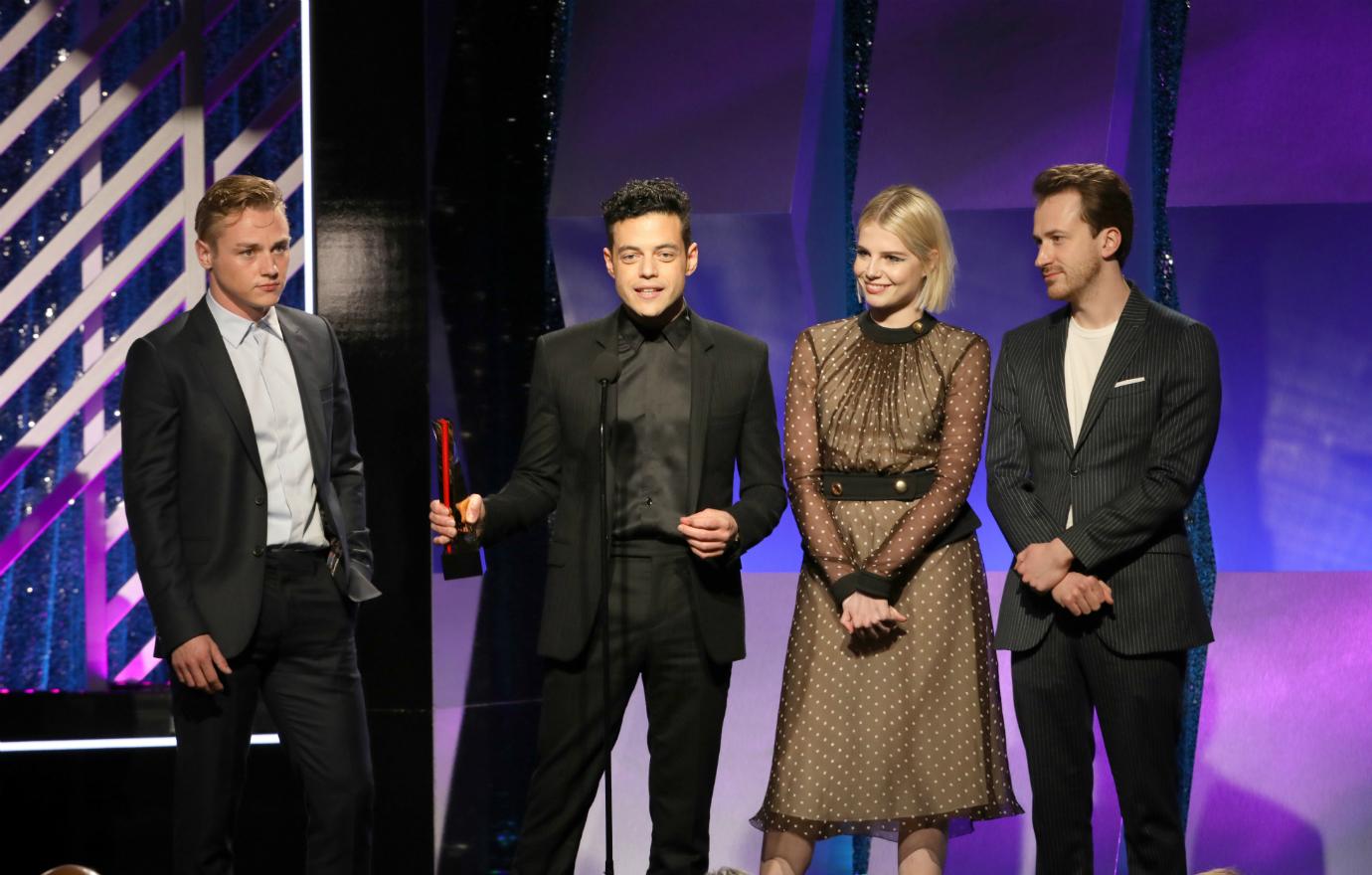 Lucy Thornton, in a gold dress, is flanked by Rami Malek and Bohemian Rhapsody stars.