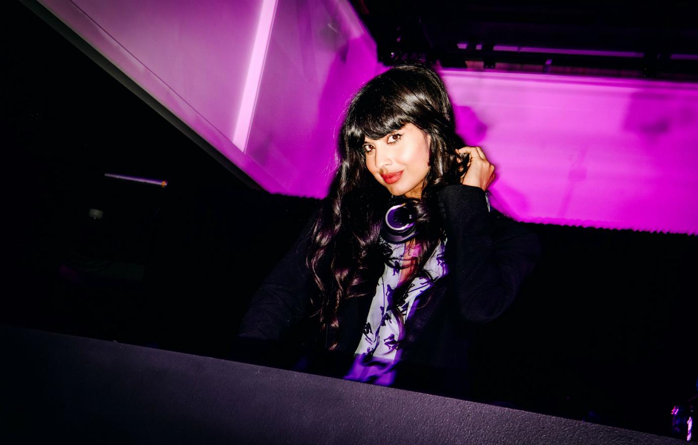 Jameela Jamil in a black jacket and purple blouse stands by the DJ deck.