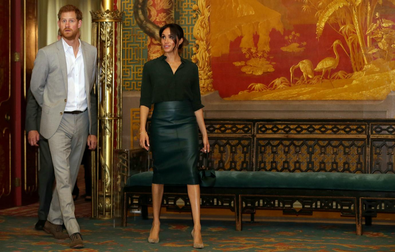 Prince Harry, in a grey suit and white shirt, walks hands clasped behind his back next to Meghan Markle who wears a black leather skirt and blouse.