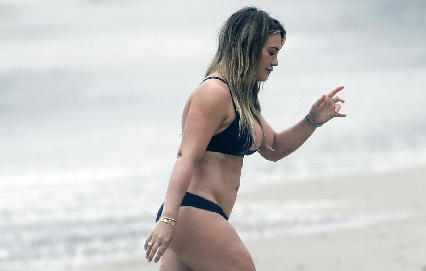 Hilary Duff, in a black bikini, walks up the beach from the shore.