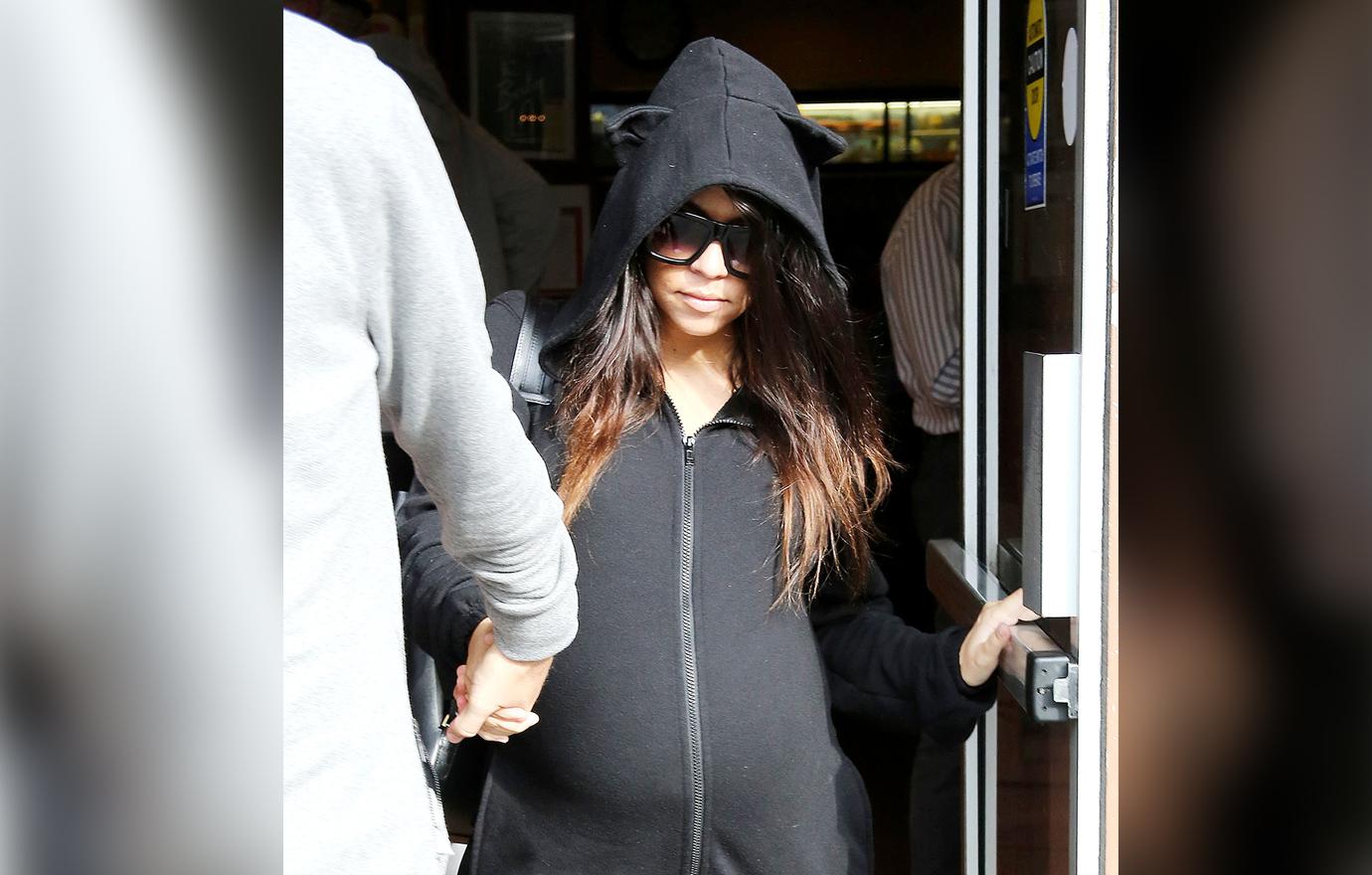 A pregnant Kourtney Kardashian in a zipped up black hoody and sunglasses exits a building.