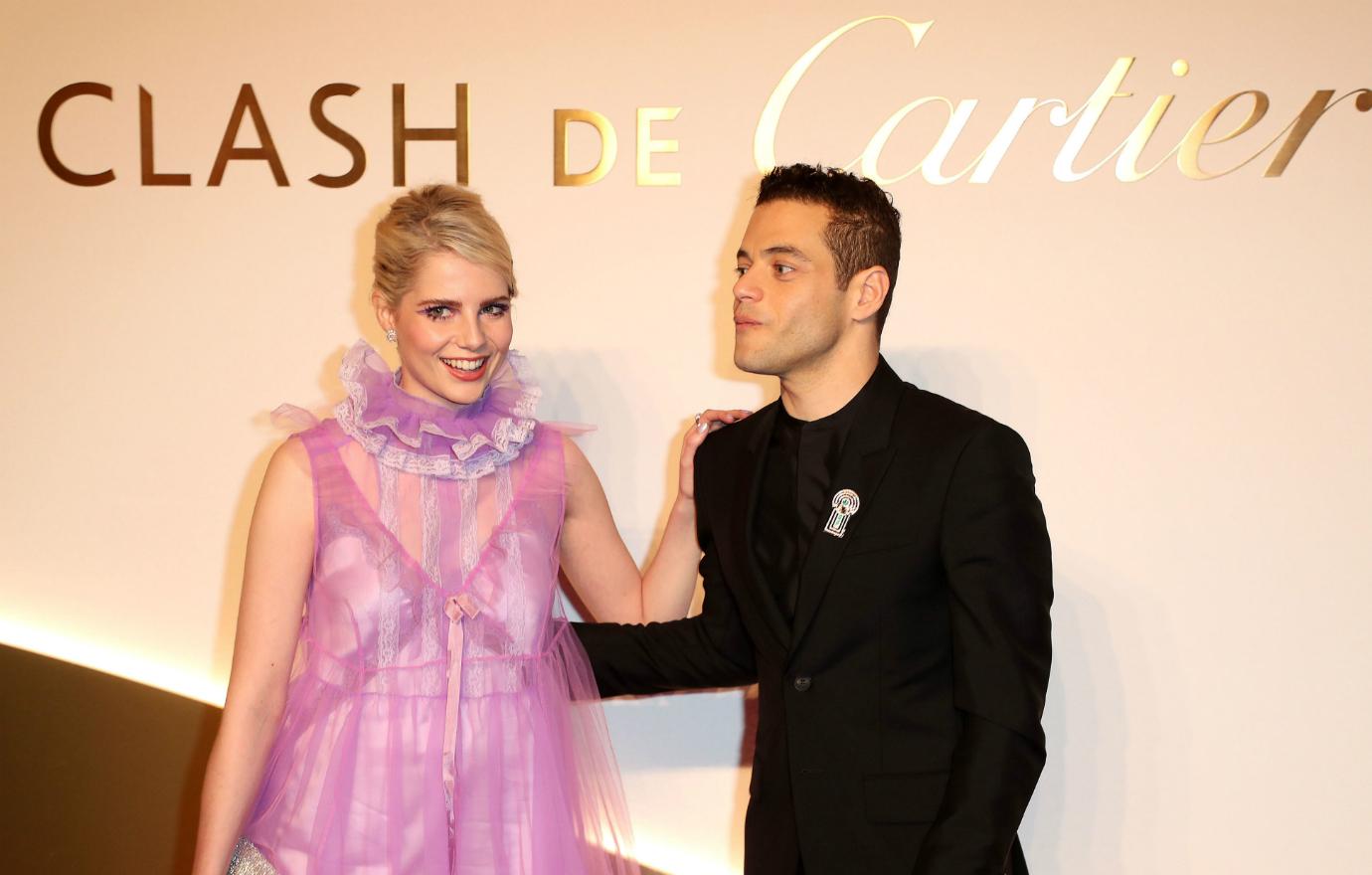 Lucy Boynton, in a pink dress stands next to Rami Malek who wears all black.