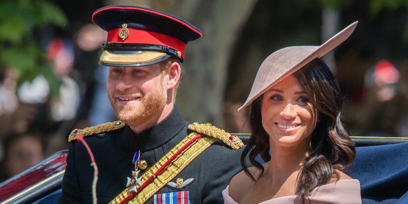 Prince Harry, in royal garb and hat, sits in a carriage with Meghan Markle who wears a matching pink dress and hat.