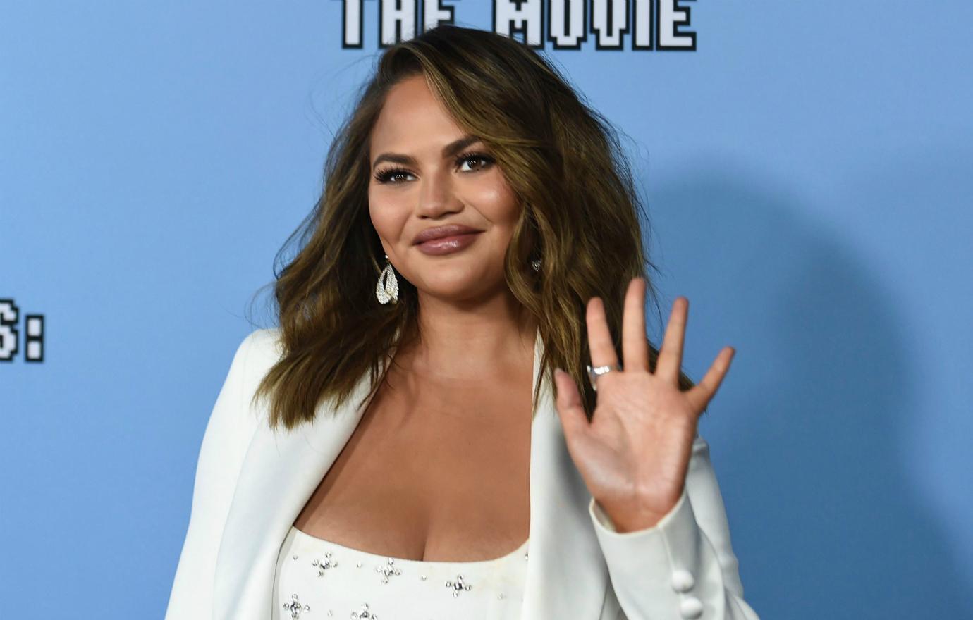 Chrissy Teigen, in a white outfit, waves at the cameras while on the red carpet.