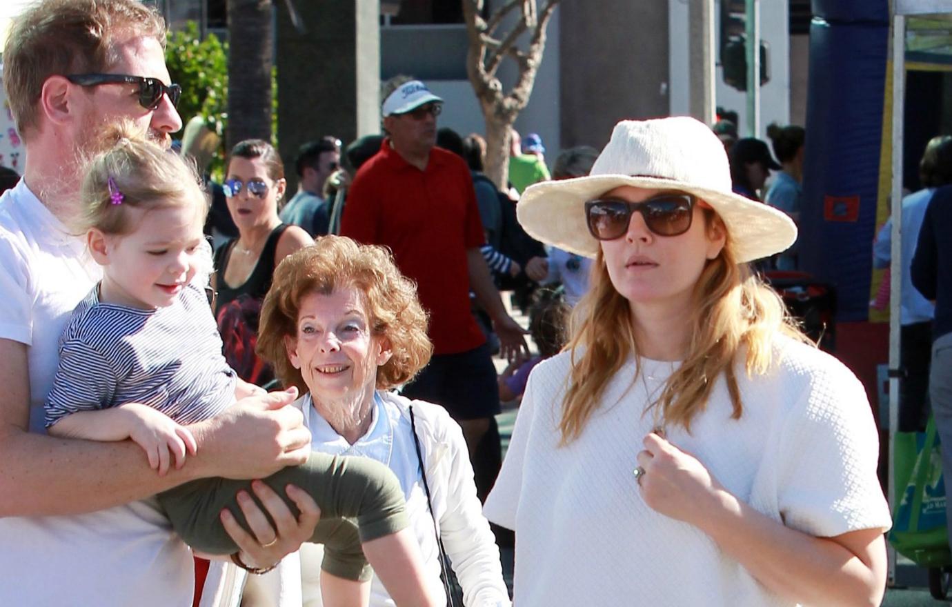 Drew Barrymoore, in sunglasses, white hat, and green jacket pushes a crib alongside Will Kopelman who wears a baseball cap and layered shirts.