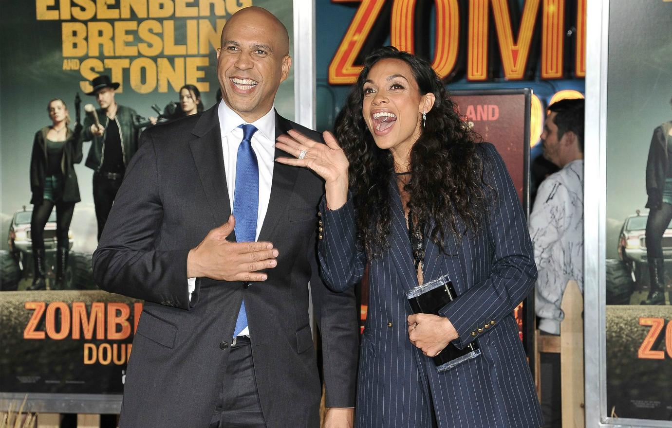 Cory Booker, in dark suit with white shirt and blue tie, points to Rosario Dawson who stands next to him smiling.