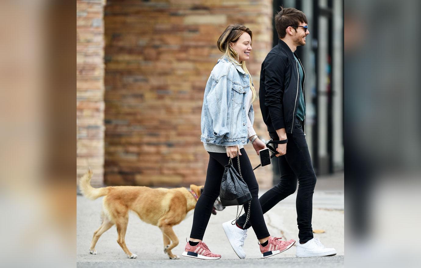 Hilary Duff, in a blue denim jacket, black jeans walks outside with Matt Koma, in black jacket and jeans and a dark green top, and their light brown dog.