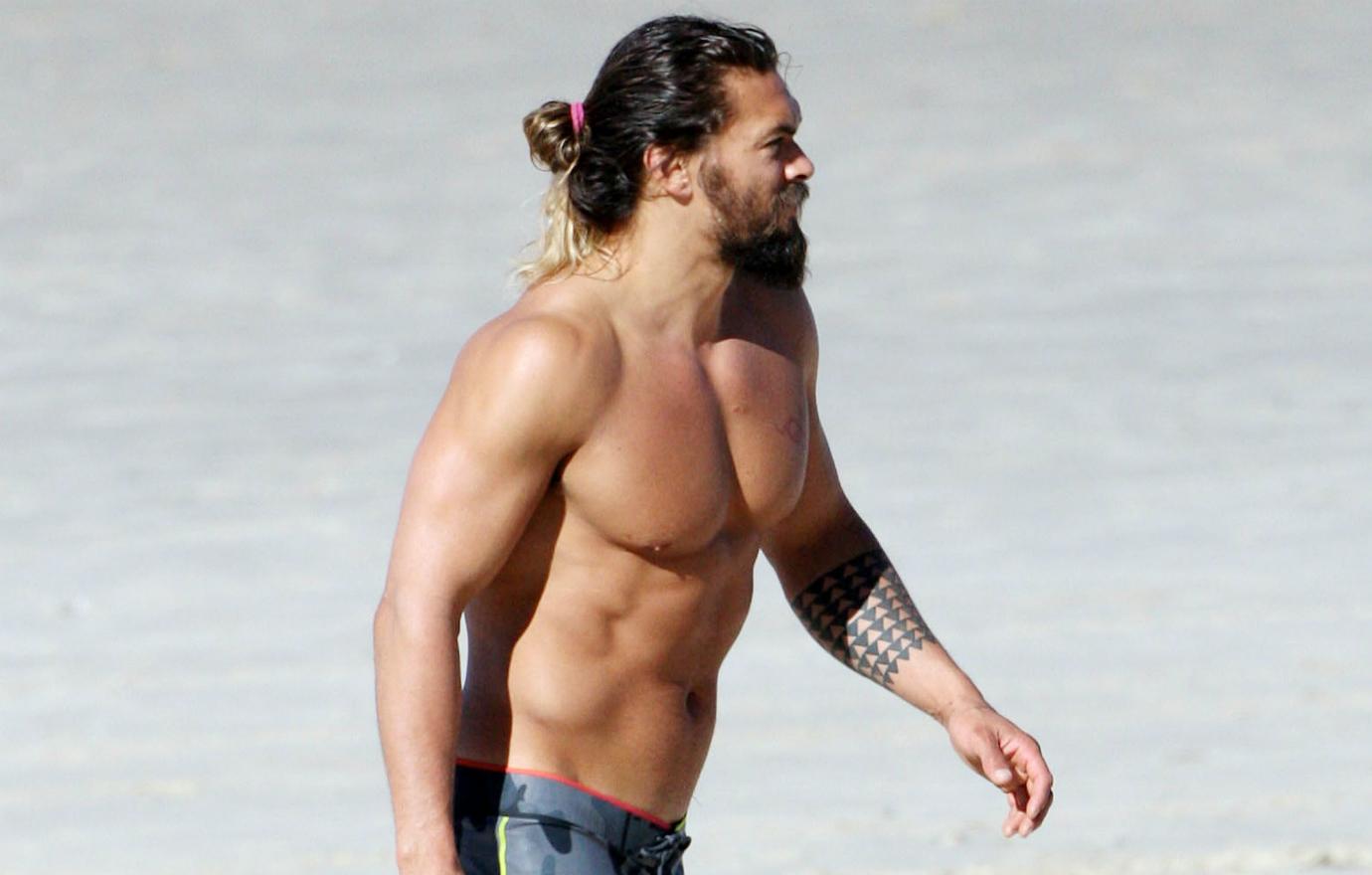 Jason Momoa walks shirtless but in shorts along a beach.