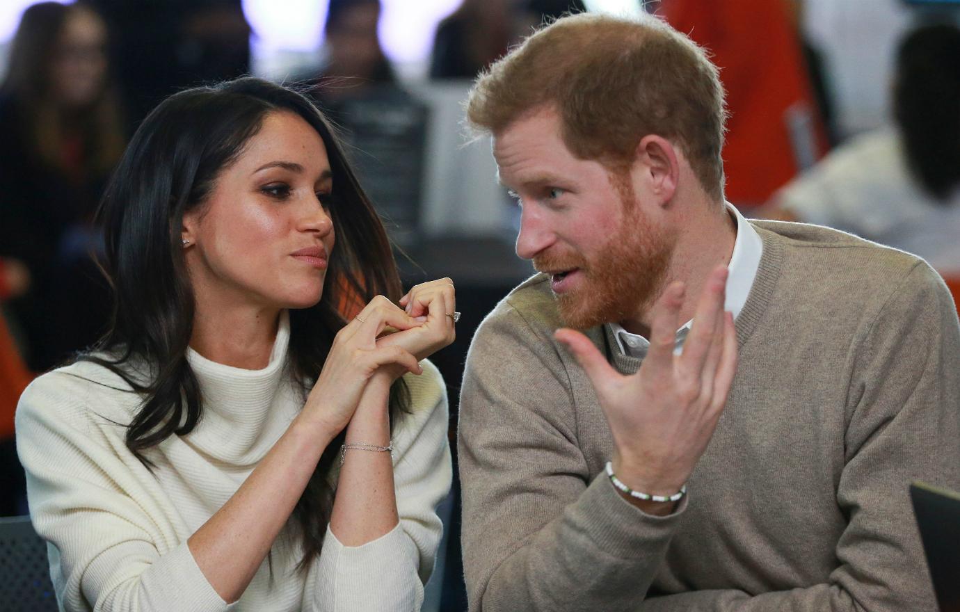 Prince Harry, in a beige sweater, talks to Meghan Markle, in a white top, who listens raptly.