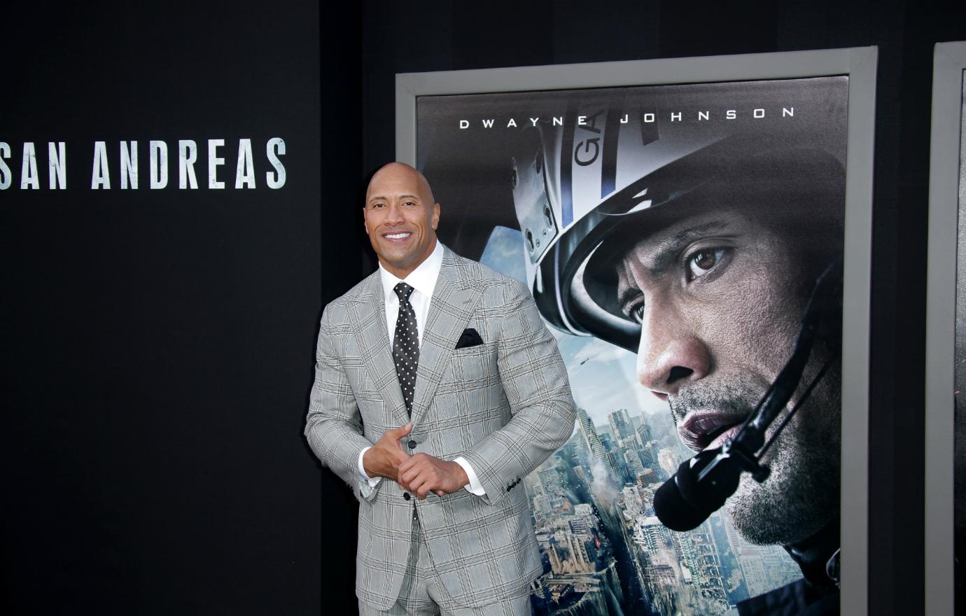 THe Rock, in a grey suit, smiles on the San Andreas red carpet event.