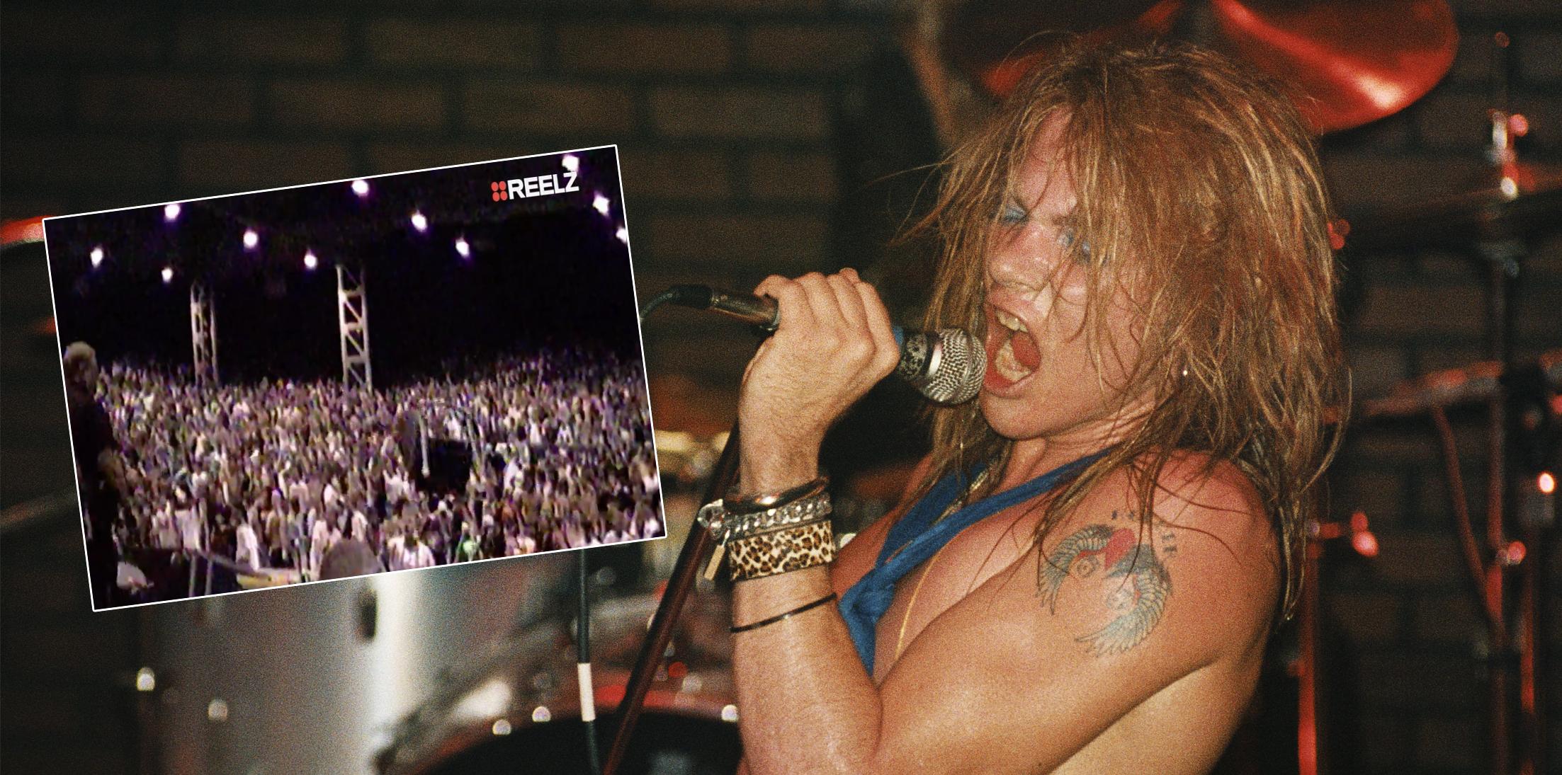 Axl Rose grips a microphone while wearing no shirt and sporting a blue scarf.