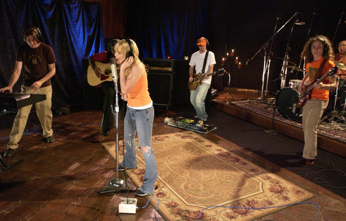 Hilary Duff wears an orange top and blue jeans and sings into a mic as her band is gathered around her on stage.